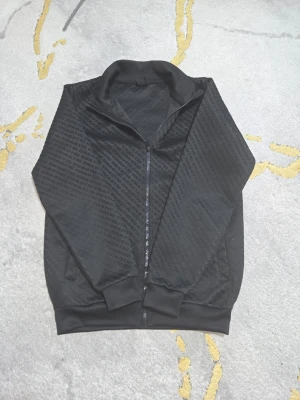 Svart zip hoodie med diskret mönster - Snygg svart hoodie med dragkedja och diskret mönster över hela tyget. Tröjan har ribbade muddar vid ärmslut och nederkant samt ståkrage. Perfekt för en stilren och avslappnad look.