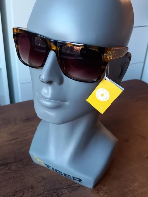 Bruna solglasögon från Sunshine  - Snygga solglasögon från Sunshine med bruna bågar och röda detaljer på skalmarna. Glasen är tonade och bågarna har en klassisk, något fyrkantig form. Perfekta för att lyfta din stil under soliga dagar.