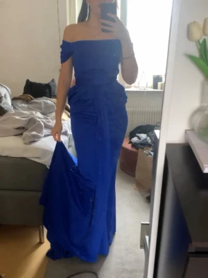 Blå axelbandslös långklänning - Elegant blå långklänning med offshoulder-design och figurnära passform. Klänningen har en markerad midja och lätt drapering vid höfterna, vilket ger en snygg siluett. Perfekt för bal eller festliga tillfällen.