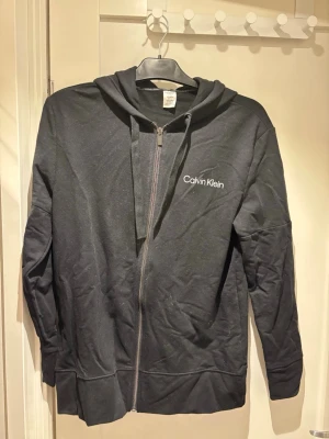 Svart zip-hoodie från Calvin Klein | S - Mysig svart loungewear-hoodie från Calvin Klein med dragkedja och huva. Diskret logga på bröstet och breda snören i huvan. Perfekt för chill dagar hemma. Tillverkad i mjukt material för extra komfort.