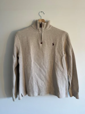 Polo Ralph Lauren - Quarter Zip - Ralph Lauren quarter-zip i klassisk oatmeal ribbed cotton, stl L. Läderzipper, broderad pony i burgundy – tidlös colorway som funkar till allt. Köpt secondhand, inget kvitto. Skick väldigt fin