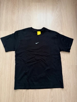 Svart NOCTA x Nike t-shirt - Svart t-shirt från NOCTA x Nike med klassisk rund hals och vit Nike Swoosh-logga på bröstet. T-shirten har en enkel och clean design, perfekt för en stilren look. Tillverkad i mjuk bomull för skön komfort.