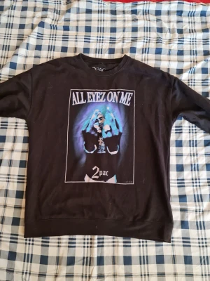 Svart sweatshirt med 2Pac-tryck - Cool svart sweatshirt med stort tryck av 2Pac och texten 'All Eyez On Me' på framsidan. Tröjan har rund hals och långa ärmar, perfekt för dig som gillar streetstyle och hiphop-vibe.