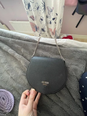 Svart handväska från Guess - Snygg svart handväska från Guess med rundad form och guldfärgad logga framtill. Väskan har en kedjerem i silver och är tillverkad i strukturerat konstläder. Magnetlås och ett litet innerfack. Perfekt för att bära det viktigaste.