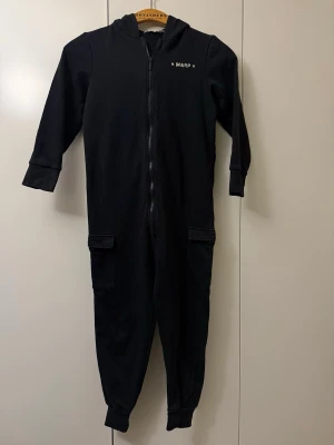 Svart onesie med huva från Warp - Svart onesie från Warp med huva och dragkedja framtill. Långa ärmar, muddar vid ärmslut och benslut samt fickor på benen. Tryck med Warp-logga på bröstet och grafiskt tryck på ryggen. Perfekt för chill hemma.
