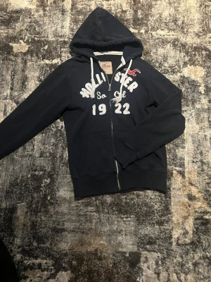 Mörkblå hoodie från Hollister - Mörkblå hoodie från Hollister med vit broderad text och siffror framtill. Klassisk huva med dragsko, hel dragkedja och två fickor. Perfekt för en avslappnad och cool stil. Är Xl men är verkligen inte det skulle säga att den passar S-M