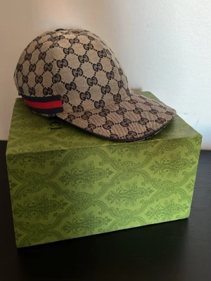 Gucci keps beige - Snygg keps från Gucci i beige canvas med klassiskt GG Supreme-mönster och röd-blå rand på sidan. Justerbar baktill och böjd skärm. Perfekt för dig som vill ha en exklusiv accessoar med streetkänsla.