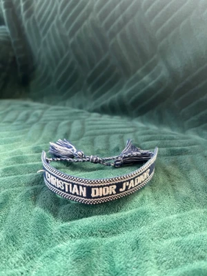 Blått broderat armband Dior - Snyggt blått armband från Christian Dior med texten 'J'ADIOR' broderad i vitt. Armbandet är gjort i tyg och har justerbara tofsar för en cool och personlig look. Perfekt accessoar för att lyfta din outfit.