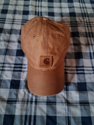 Brun keps från Carhartt - Snygg brun keps från Carhartt i klassisk modell med böjd skärm. Kepsen är tillverkad i 100% bomull och har justerbar passform. Perfekt accessoar för en avslappnad streetwear-look.