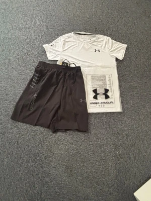 Under Armour träningsset  - Säljer ett fräscht träningsset från Under Armour med en vit t-shirt och svarta shorts. T-shirten har tryck på bröstet, ärmen och sidan, och shortsen har elastisk midja och logga på benet. Perfekt för gym eller löpning. Står M fast passar M/S