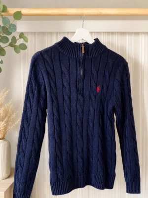 Mörkblå kabelstickad tröja Polo Ralph Lauren - Kabelstickad mörkblå tröja från Polo Ralph Lauren med halv dragkedja och ribbad krage. Tröjan har lång ärm och röd broderad logga på bröstet. Perfekt för lager-på-lager och snygg till jeans eller linnebyxor. LAPP FINNS KVAR