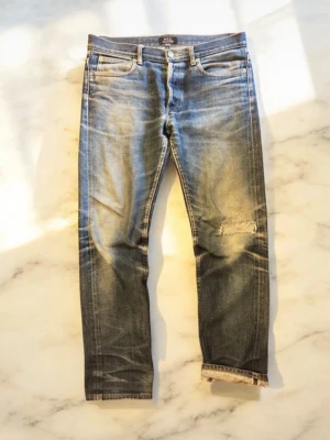 A.P.C. Selvage jeans - Säljer ett par blå jeans från A.P.C. med snyggt slitna detaljer och patch på ena knät. Jeansen har klassisk femficksmodell, raka ben och tydliga slitningar för en cool vintagekänsla. Perfekta för dig som gillar streetwear och avslappnad stil.