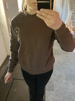 Brun sweatshirt med tryck - Säljer en brun sweatshirt med rund hals och tryck på vänster bröst. Tröjan har ribbade muddar vid ärmslut och nederkant. Perfekt för en avslappnad och stilren look. Materialet känns mjukt och bekvämt mot huden.