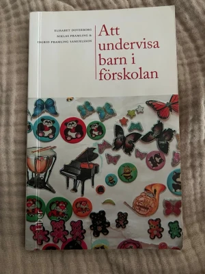 Att undervisa barn i förskolan - Boken 'Att undervisa barn i förskolan' av Elisabet Doverborg, Niklas Pramling och Ingrid Pramling Samuelsson tar upp konkreta principer för undervisning i förskolan. Omslaget är vitt med färgglada klistermärken av djur, instrument och fjärilar.