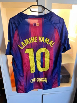 FC Barcelona Lamine Yamal matchtröja - Säljer en officiell FC Barcelona matchtröja från Nike med Lamine Yamal och nummer 10 på ryggen. Tröjan är blå och vinröd med gula detaljer, klubbmärke och sponsorloggor framtill. Tillverkad i lätt och ventilerande funktionsmaterial, perfekt för fotbollsträning.