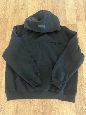 Svart Essentials Fear of God hoodie - Svart hoodie från Essentials Fear of God med diskret logga på bröstet. Klassisk passform med huva och känguruficka framtill. Tillverkad i mjukt bomullsmaterial som ger en skön och avslappnad känsla.