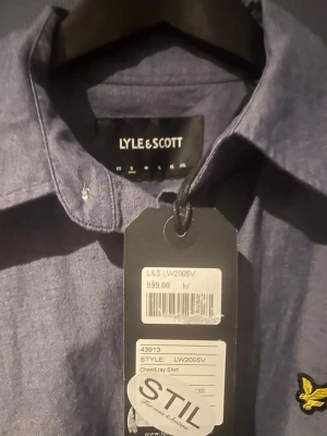 Blå skjorta från Lyle & Scott - Stilren blå skjorta från Lyle & Scott med klassisk krage och knäppning framtill. Skjortan har lång ärm och den ikoniska gula loggan broderad på bröstet. Perfekt för en clean och snygg look.