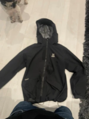 Svart vindjacka från Haglöfs - Svart vindjacka från Haglöfs med huva och hel dragkedja framtill. Jackan har två sidofickor med dragkedja och reflekterande detaljer på ärmarna. Perfekt för blåsiga dagar och outdoor-aktiviteter. Lätt material och sportig design. Men det finns ett litet hål i luven annars så är den topp skick