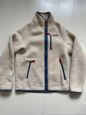 Beige fleecejacka från Patagonia - Säljer en beige fleecejacka från Patagonia med blå detaljer och röda dragkedjor. Jackan har hög krage, tre fickor med dragkedja och är tillverkad i mjukt teddyfleece-material. Perfekt för lager-på-lager och outdoor vibes.