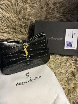 Svart croco handväska  - Snygg svart handväska i croco-präglat läder med guldigt logo framtill. Kommer med dustbag och originalkartong. Perfekt accessoar för dig som vill ha en lyxig och trendig look. 