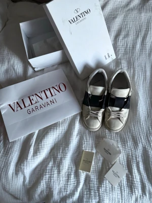 Valentino Garavani  - Snygga vita sneakers från Valentino Garavani med bred svart rem över vristen. Klassisk låg modell med rund tå, vita skosnören och diskret logga på plösen. Lyxig känsla och stilren look.