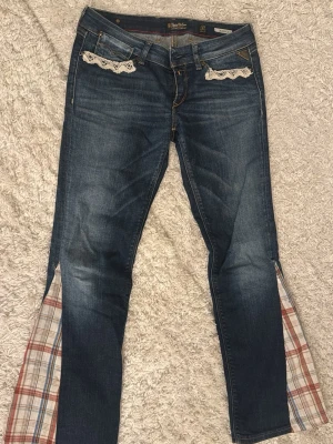 Unika jeans med rutiga detaljer - Mörkblå jeans med rutiga tygkilar i benen och hjärtformade rutiga detaljer på bakfickorna. Fickorna fram har vit spets som dekoration. Jeansen har bootcut-modell och cool vintagekänsla. Perfekta för dig som vill sticka ut med din stil.