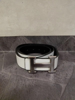 Vitt bälte - Snyggt vitt läderbälte  med ett stort, silverfärgat H-spänne. Bältet har en klassisk design med svarta kanter och flera hål för justerbar passform. Perfekt accessoar för att lyfta din outfit.