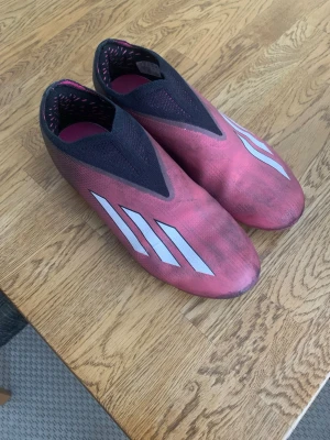 Rosa Adidas fotbollsskor utan snörning - Säljer ett par rosa Adidas fotbollsskor med vita stripes på sidan och svart mesh vid öppningen. Skorna har en strömlinjeformad design utan snörning och fasta dobbar för bra grepp på planen. Perfekta för dig som vill ha stilrena och snabba skor på matchen.