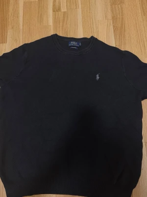Svart tröja från Polo Ralph Lauren - Klassisk svart tröja från Polo Ralph Lauren med rund halsringning och diskret broderad logga på bröstet. Tröjan har ribbade muddar vid ärmslut och nederkant. Perfekt för en stilren och enkel look.