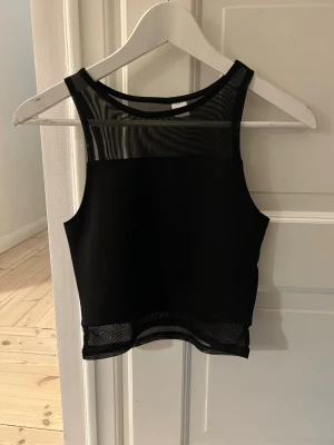 Svart crop top med meshdetaljer - Snygg svart crop top med transparenta meshpartier upptill och nedtill. Ärmlös modell med rund halsringning och figurnära passform. Perfekt att styla till höga jeans eller kjol för en trendig look.