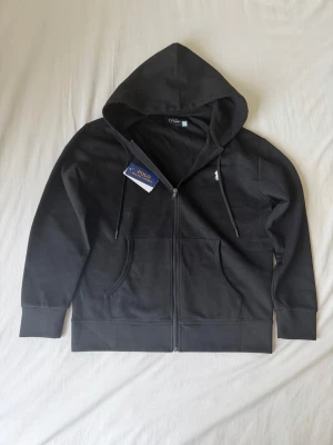 Svart zip hoodie från Polo Ralph Lauren - Snygg svart hoodie med dragkedja från Polo Ralph Lauren. Klassisk design med huva och dragsnören, två fickor framtill och diskret logga på bröstet. Tillverkad i mjuk bomullsblandning som känns skön mot huden. Perfekt för en avslappnad och stilren look. Storlek S