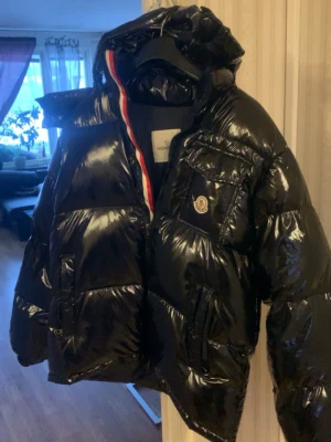 Svart glansig pufferjacka Moncler - Säljer en svart glansig pufferjacka från Moncler med huva och flera fickor framtill. Jackan har en ikonisk Moncler-logga på bröstet och röd-vit-blå detalj på insidan av dragkedjan. Tillverkad i blankt material som ger en riktigt lyxig känsla.