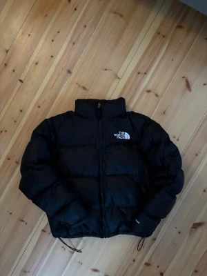 The North Face 1996 Retro Nuptse Jacket - Säljer en svart dunjacka från The North Face, modell 700. Jackan har hög krage, dragkedja framtill och logga på bröstet. Puffig design med dragsko i nederkant och broderad 700-detalj på ärmen. Perfekt för kalla vinterdagar. Detta är den klassiska, trendiga North Face pufferjacka som trendar på tiktok.