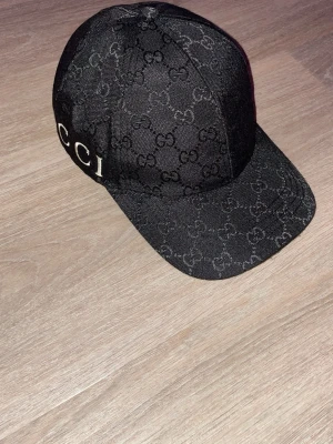 Svart Gucci keps med monogram - Snygg svart keps från Gucci med klassiskt GG-monogram över hela kepsen och broderad logga runt bakre delen. Justerbar rem i läder baktill för perfekt passform. Perfekt accessoar för dig som gillar exklusiv streetwear och vill sticka ut.
