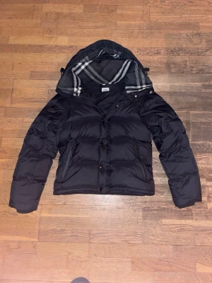 Svart burberry dunjacka med huva i storlek s men avtagbara ärmar - Snygg svart dunjacka med avtagbar huva och rutigt foder i grått och vitt. Jackan har knappar och dragkedja framtill samt två sidofickor. Puffig modell som håller dig varm under kalla dagar. Perfekt för vinter och har en klassisk look.