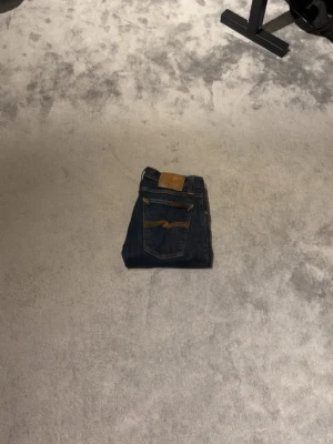 Mörkblå jeans från Nudie Jeans - Säljer ett par mörkblå jeans från Nudie Jeans med klassisk femficksdesign och kontrastsömmar på bakfickorna. Jeansen har en läderpatch med logga bak i midjan och är tillverkade i kraftigt denimtyg. Perfekta för en avslappnad och stilren look. Storleken är W27 L34 