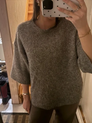 Grå stickad tröja med halvlånga ärmar - Mysig grå stickad tröja med rund halsringning och halvlånga, vida ärmar. Tröjan har en avslappnad passform och ribbstickade kanter vid hals, ärmslut och nederkant. Perfekt för lager på lager och enkel att styla till olika outfits.