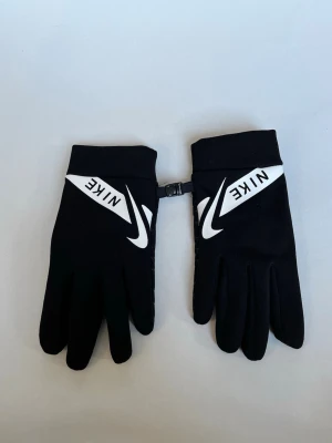 Svarta sporthandskar från Nike - Snygga svarta handskar från Nike med vit logga och reflexdetaljer. Handskarna har greppmönster i handflatan och touchfunktion på pekfingret. Perfekta för träning eller vardag när du vill hålla händerna varma och ändå kunna använda mobilen.