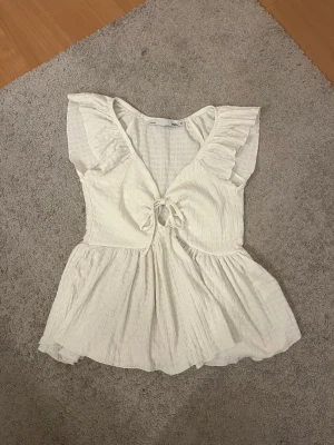 Vit volangtopp med knyt XS - Supersöt vit topp från lager 157 i storlek XS. Toppen har volangdetaljer vid axlarna, v-ringning och knyt framtill. Tillverkad i ett lätt, strukturerat bomullstyg som ger en luftig känsla. Perfekt för varma dagar och en romantisk look.