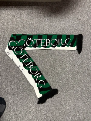 Grönsvart halsduk Gais - Snygg halsduk med gröna och svarta ränder och texten 'Göteborg' samt 'Grönsvart'. Halsduken har svarta fransar i ändarna och är perfekt för att visa ditt stöd för laget. Materialet är stickat tyg. Och signerat av 5 Gais spelare kommer ihåg att en av dom var Axel Henrikson, 