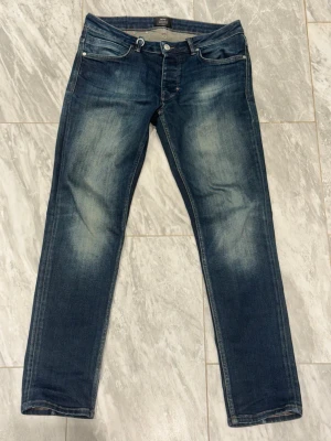 Mörkblå slim fit jeans med slitningar - Snygga mörkblå slim fit jeans med slitna detaljer och ljusa tvättade partier på framsidan och baksidan. Klassisk femficksmodell med knappgylf och raka ben. Perfekta för en avslappnad och trendig look.