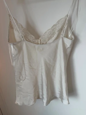 Vit satinlinne med spetsdetaljer - Säljer ett elegant vitt linne i satin med tunna axelband och vacker spets längs urringningen. Perfekt för en chill dag hemma eller som ett lager under en kofta. Materialet känns mjukt mot huden och ger en lyxig känsla.
