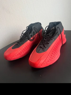 Anthony Edwards 1 adidas - Säljer ett par riktigt snygga Adidas sneakers i rött och svart. Skorna har en futuristisk design med mesh- och syntetmaterial, samt tydliga Adidas-loggor på hälen. De har svarta snören och en cool struktur på sidorna som ger extra edge.