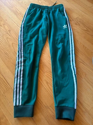 Gröna Adidas mjukisbyxor med ränder - Säljer ett par gröna mjukisbyxor från Adidas med klassiska vita ränder längs benen och Adidas-logga på vänster sida. Byxorna har resår i midjan och muddar vid bensluten. Perfekta för chill eller träning, riktigt sköna och stilrena.