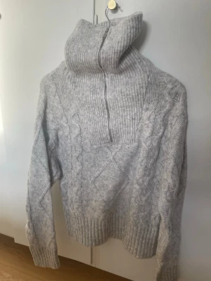 Ljusgrå stickad polotröja med dragkedja - Säljer en ljusgrå stickad tröja med hög polokrage och dragkedja framtill. Tröjan har ett klassiskt flätmönster längs ärmar och framstycke samt breda ribbade muddar. Perfekt för kalla dagar och riktigt mysig att bära. 💕