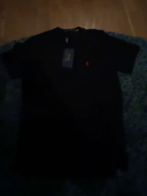 Svart t-shirt från Polo Ralph Lauren - Klassisk svart t-shirt från Polo Ralph Lauren med röd broderad logga på bröstet. T-shirten har rund halsringning och korta ärmar. Tillverkad i mjuk bomull för en skön känsla och enkel stil.