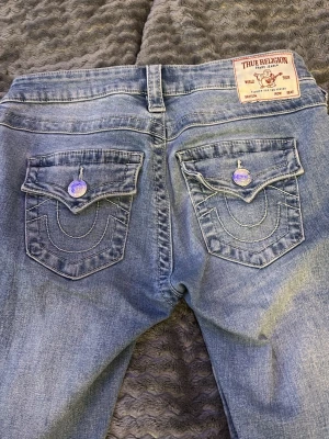 Blå bootcut jeans från True Religion - Säljer ett par blå bootcut jeans från True Religion med ikoniska kontrastsömmar och stora fickor bak med lock och dekorativa knappar. Jeansen har en klassisk tvättad look och är tillverkade i denim med snyggt fall.