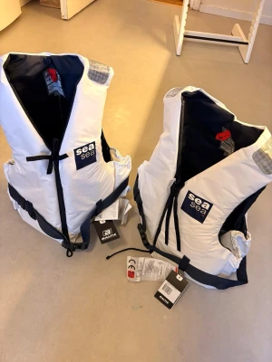 Vita flytvästar från SeaSea - Två vita flytvästar från SeaSea med svarta detaljer och reflexer på axlarna. Västarna har justerbara remmar i midjan och snörning nedtill. Märket syns tydligt framtill och materialet är slitstark syntet. Perfekt för båtliv och vattensport.