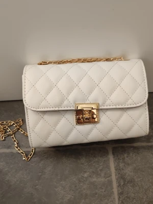 Vit quiltad axelväska med guldkedja - Snygg vit axelväska med quiltat mönster och guldfärgad kedjerem. Väskan har ett elegant vridlås i guld på framsidan och en klassisk rektangulär form. Perfekt för dig som gillar stilrena accessoarer med lyxig känsla.