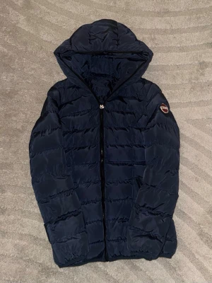 Mörkblå pufferjacka från Colmar - Snygg mörkblå pufferjacka från Moncler med huva och dragkedja framtill. Jackan har ett quiltat mönster och Moncler-logga på ärmen. Perfekt för kalla dagar och tillverkad i ett glansigt, vattenavvisande material.